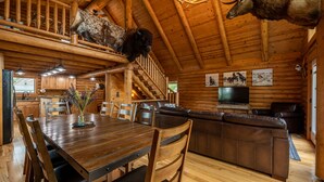 A rustic cabin interior with a wooden dining table, chairs, a sofa, and a television. - Majestic Wyoming Landscape. Riverfront log cabin. Relax in or explore out! (Meeteetse)