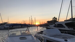 Uma marina com vários barcos atracados, um pôr do sol ao fundo e uma palmeira à vista.