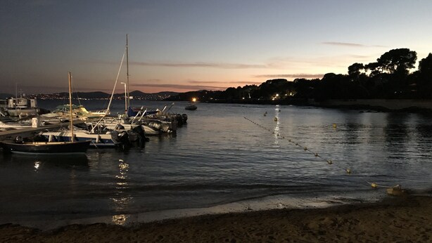 Uma marina com barcos atracados à noite, água calma e uma praia em primeiro plano.