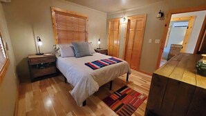 Un dormitorio con cama, mesitas de noche, lámpara, ventana con persianas, armario de madera y suelo de madera.