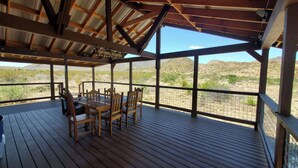 Un patio techado al aire libre con una mesa y sillas de madera, ventanas amplias y vistas al paisaje desértico.