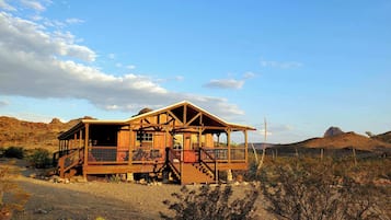 Une cabane en bois dotée d’une véranda, située dans un paysage désertique avec des montagnes en arrière-plan.
