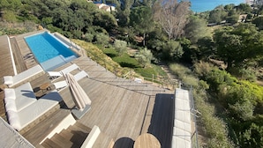 Un espace piscine avec vue sur la mer, entouré d’arbres et doté d’une terrasse en bois.