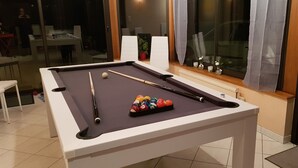 Una mesa de billar con bolas y tacos, una ventana con cortinas y una vista de un perro afuera.