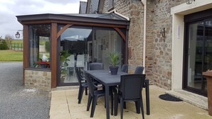 Un patio cubierto con una mesa, sillas, una planta en maceta y un edificio de piedra con puerta de cristal.
