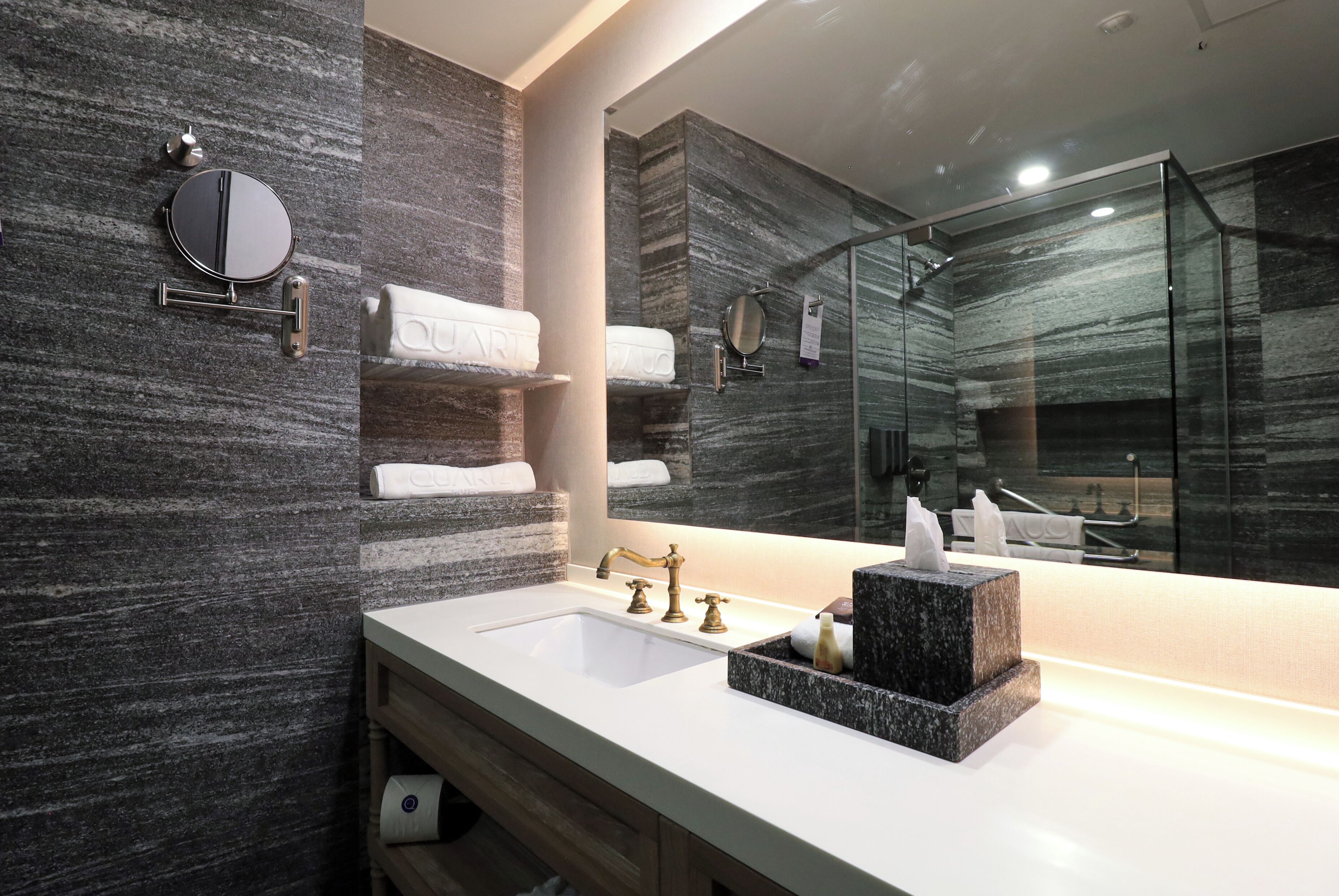 Un bagno moderno con rivestimento in marmo, un ampio specchio, un lavabo su un piano in marmo e un box doccia in vetro.