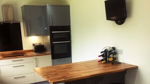 A modern kitchen with a wooden island, two stools, a built-in oven, and a wall-mounted TV. - Yorkshire Wolds Spacious Modern Holiday Bungalow (Driffield)