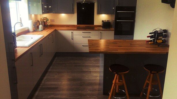 A modern kitchen with dark cabinets, a wooden countertop, a built-in oven, and two stools. - Yorkshire Wolds Spacious Modern Holiday Bungalow (Driffield)