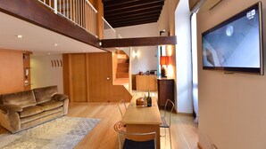 A modern living room with a wooden ceiling, a sofa, a dining table with chairs, a TV mounted on the wall, and a staircase leading to an upper level. - IzzHome Full Center Grand (Cagliari)