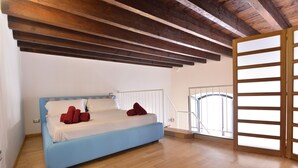 A bedroom with a blue bed, white bedding, and red pillows. It features wooden flooring, a white railing, and a view through a large window. - IzzHome Full Center Grand (Cagliari)