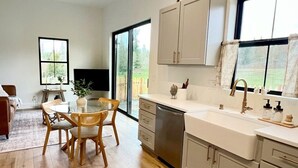 A modern kitchen with a dining area, wooden flooring, and large windows offering a view of the outdoors.