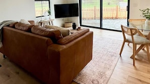 A spacious living room with a brown leather sofa, a flat-screen TV, and a dining area with wooden chairs.