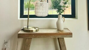 A wooden table with a lamp and a vase by a window with a view of trees and a fence.