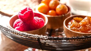 En træbakke med skåle der indeholder hindbær, appelsiner og et marmelade.