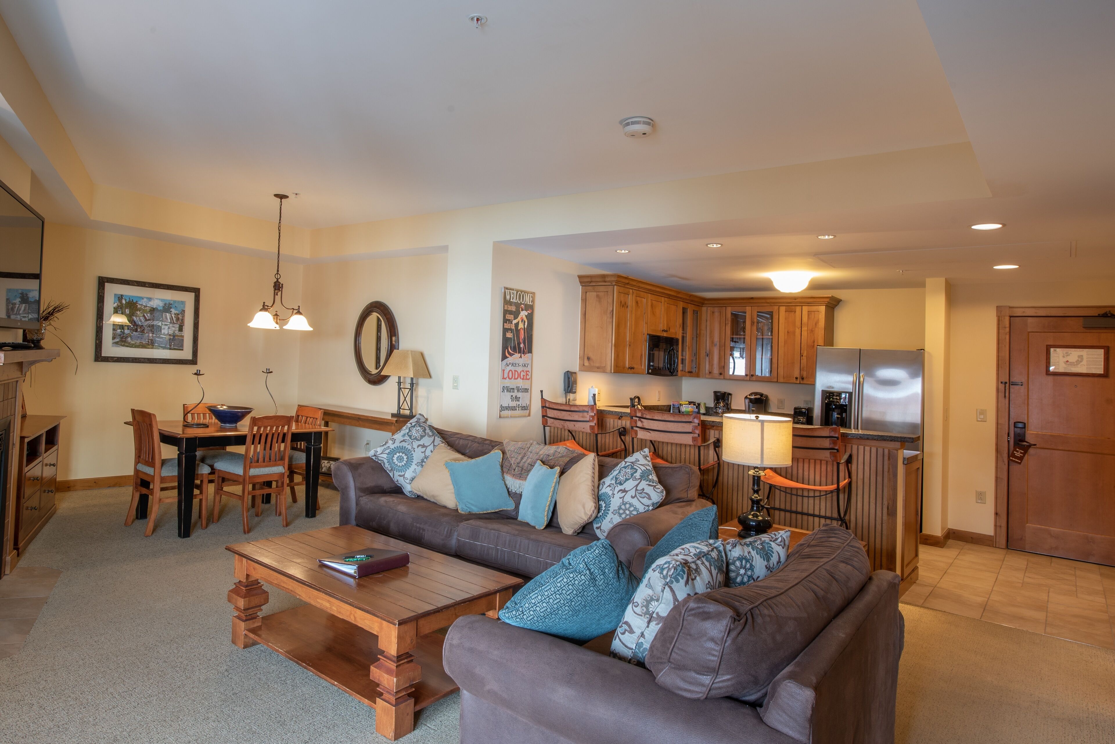 A spacious living room with a brown sofa, wooden coffee table, and a dining area with a wooden table and chairs. A kitchen with wooden cabinets and a refrigerator is visible in the background.