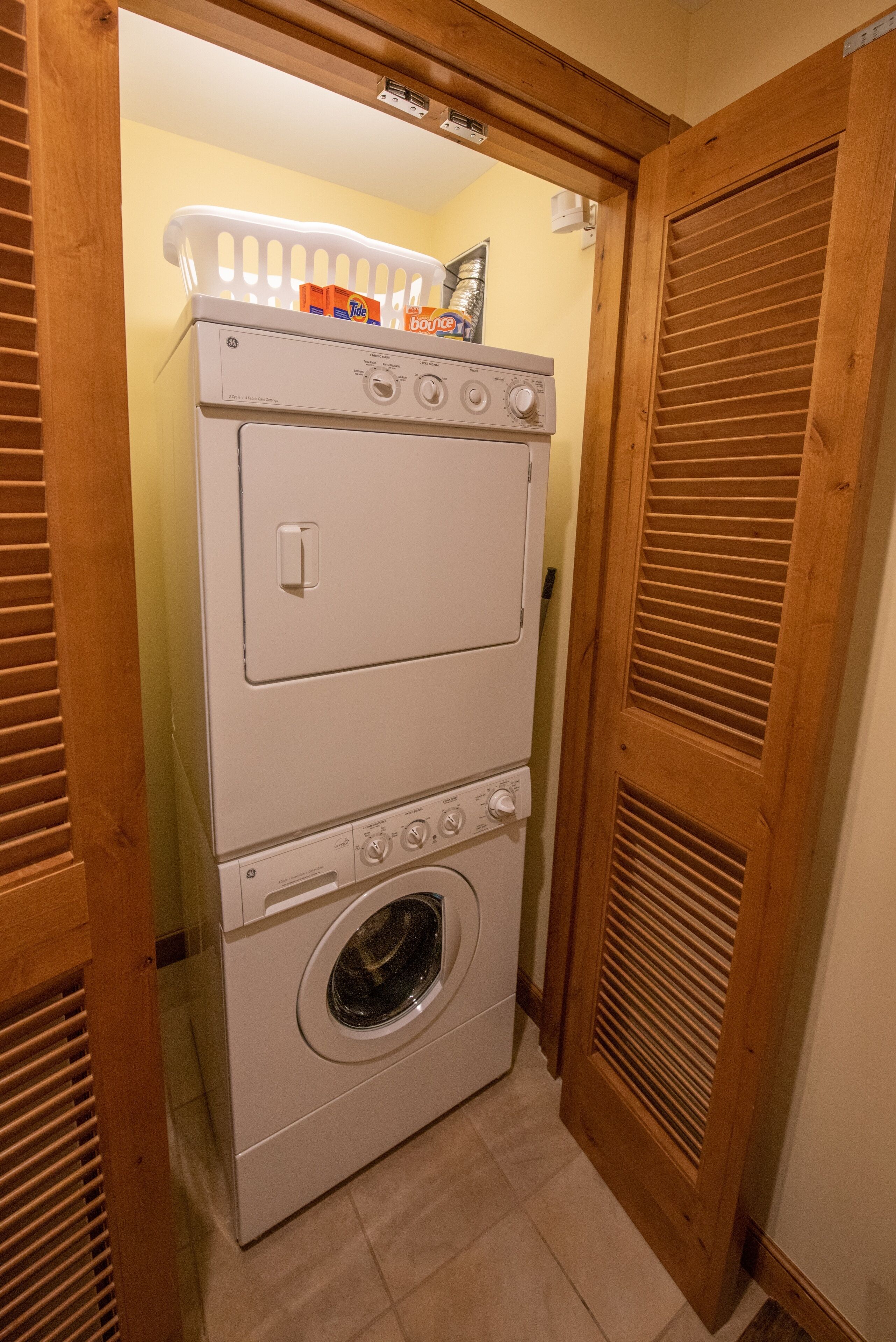 Une buanderie équipée de machines à laver et de sèche-linge superposés, d’un panier rempli de lessive et de portes en bois.