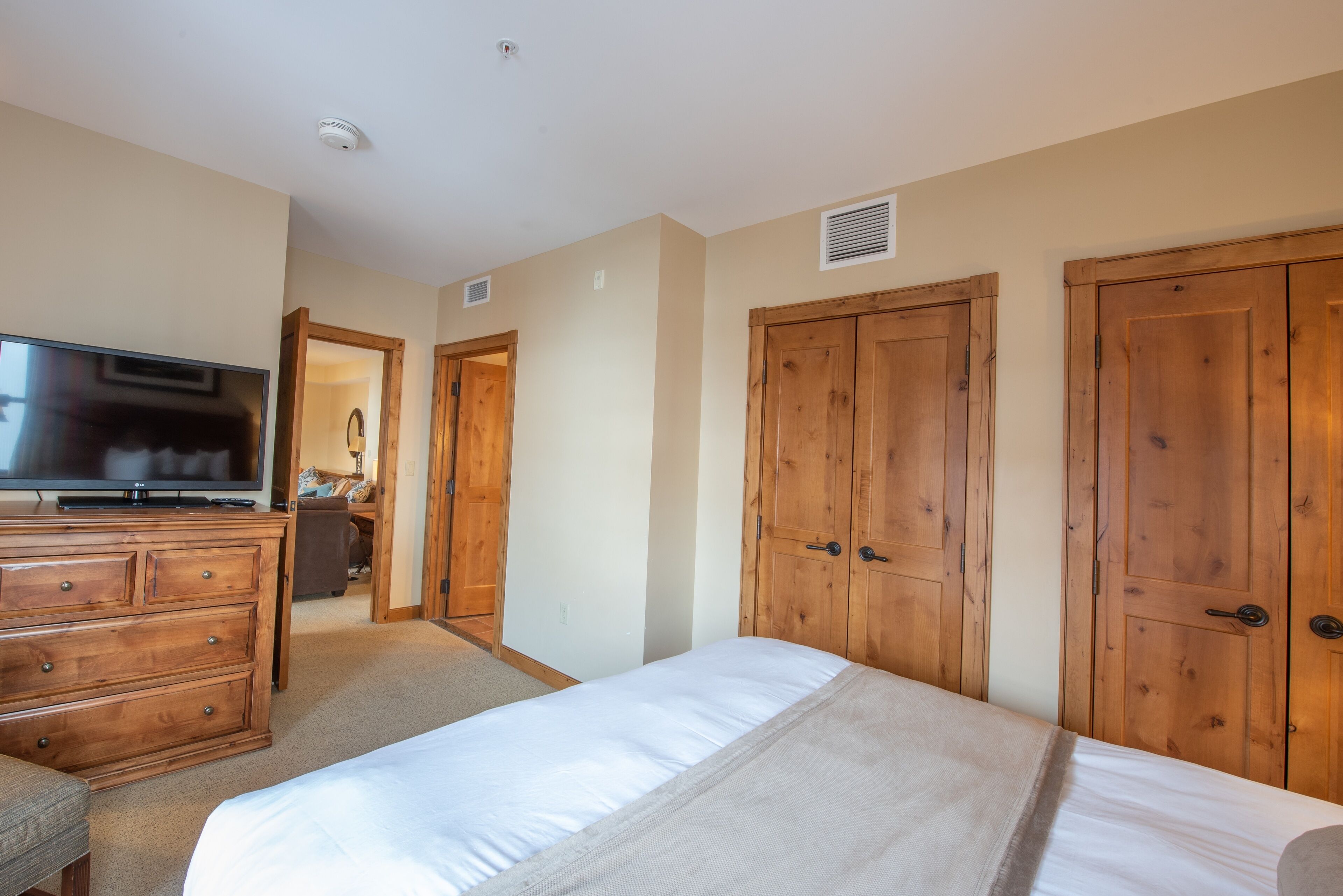A bedroom with a bed, wooden wardrobe, a TV on a dresser, and a door leading to another room.