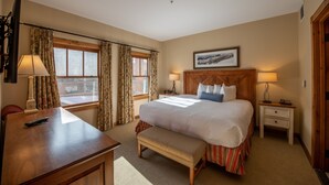 Une chambre d’hôtel avec un grand lit, un bureau en bois, un banc, une table de chevet avec une lampe et une fenêtre avec des rideaux.
