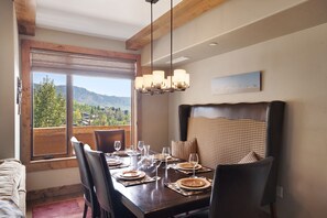 Comedor con una mesa de madera dispuesta para cuatro, vista a zonas verdes y colinas a través de grandes ventanales, y una lámpara de techo moderna.