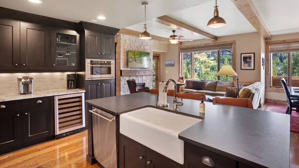 A modern kitchen with dark cabinets, a large island with a sink, and a dining area with a view of the outdoors. - Edgemont 2508 Luxury On Mountain Condo With Ski-In/Ski-Out Access! (Steamboat Springs)