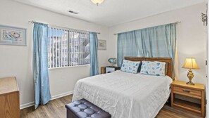 A bedroom with a large bed, a bedside table with a lamp, a bench, and curtains on the windows. - Just steps away from the beach!  (Myrtle Beach)