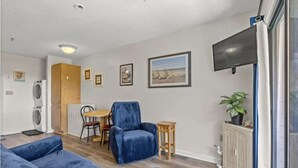 A modern living room with a blue sofa, a blue armchair, and a dining area with a table and chairs. There is a TV mounted on the wall, a plant on a cabinet, and a window with a curtain. - Just steps away from the beach!  (Myrtle Beach)