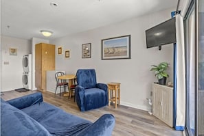 A modern living room with a blue sofa, a blue armchair, and a dining area with a table and chairs. There is a TV mounted on the wall, a plant on a cabinet, and a window with a curtain.