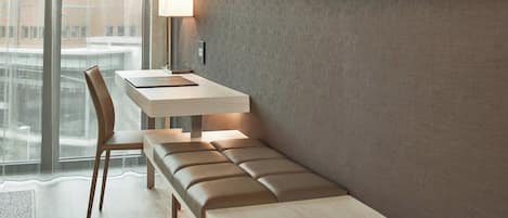A modern hotel room with a wooden desk, a cushioned bench, and a large window offering a city view.