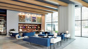 A modern hotel lobby with a blue sofa area, a bookshelf, and a large window offering a city view.
