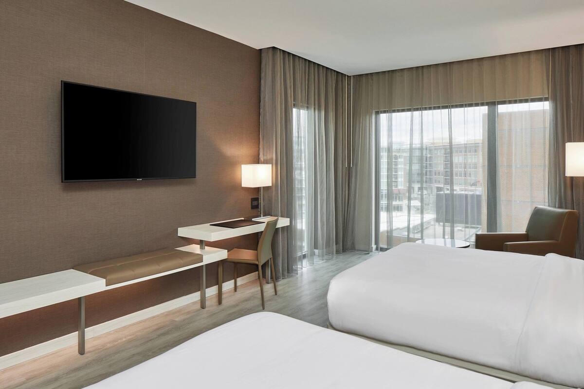 Une chambre d’hôtel moderne dotée d’une télévision à écran plat, d’un bureau avec une chaise, d’un grand lit et offrant une vue sur la ville grâce à de grandes fenêtres.