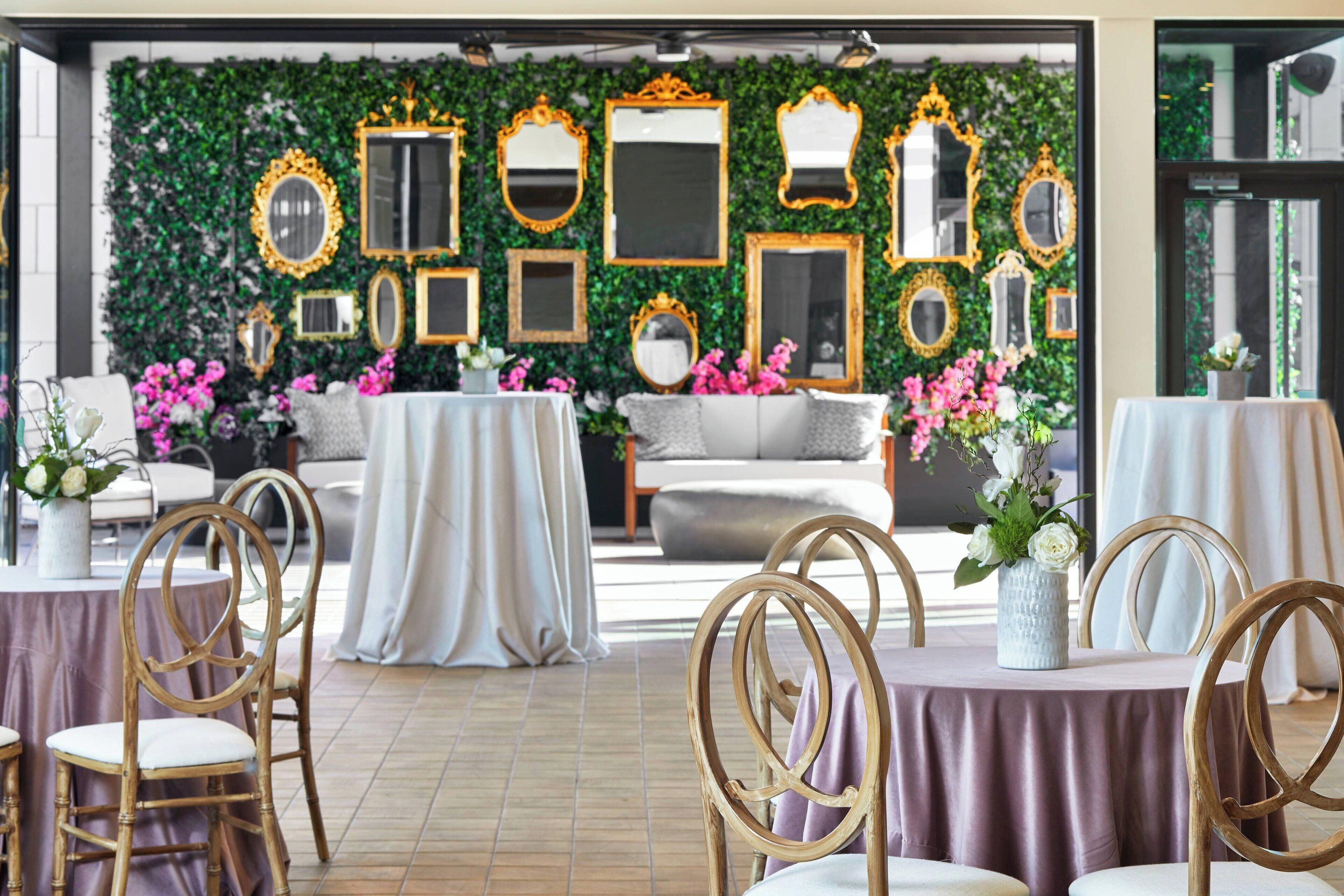 A dining area with round tables and chairs, a sofa, and a wall decorated with mirrors and greenery.