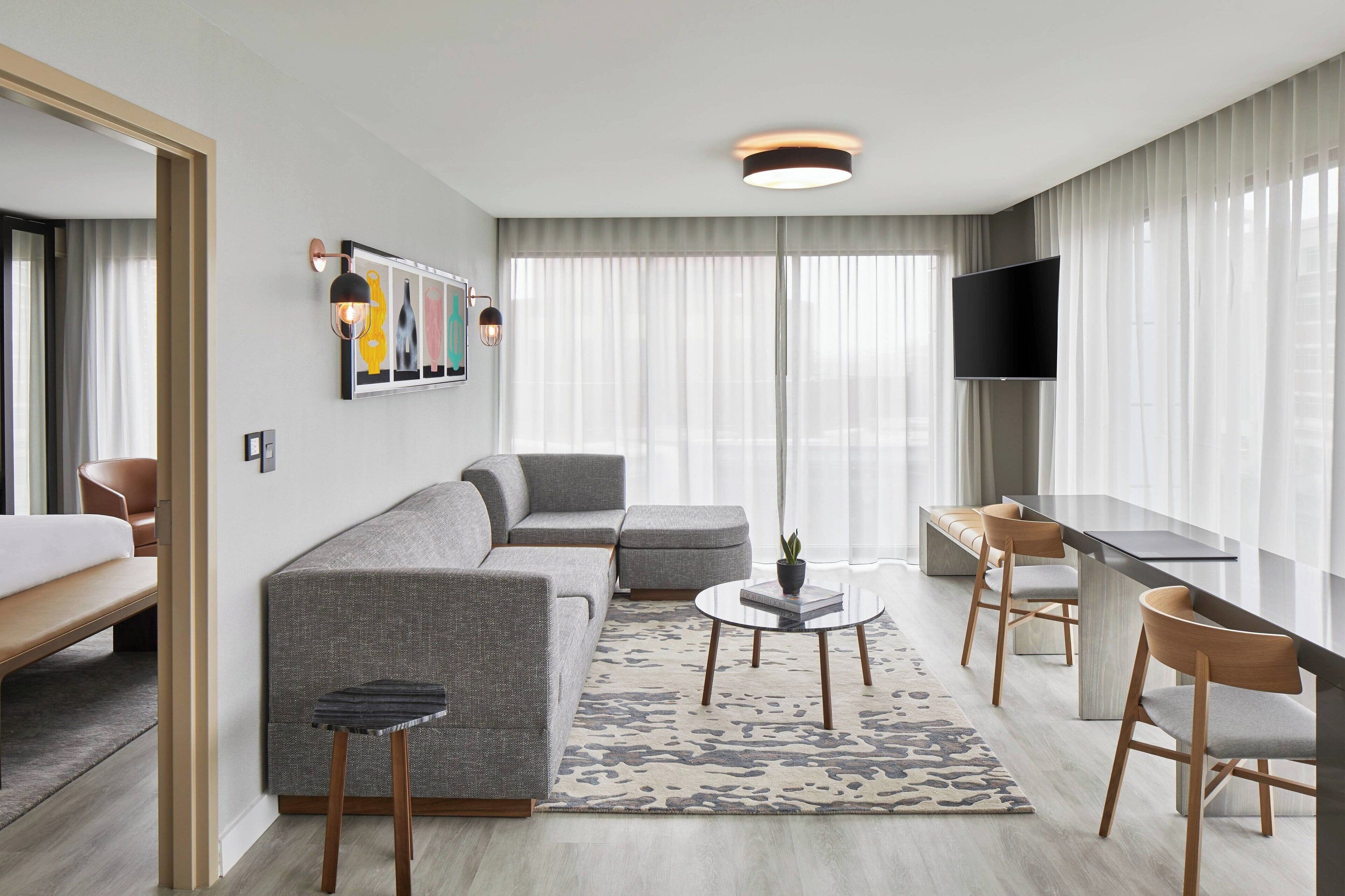 A modern hotel room with a grey sofa, a small round table, a TV, and a desk with chairs.