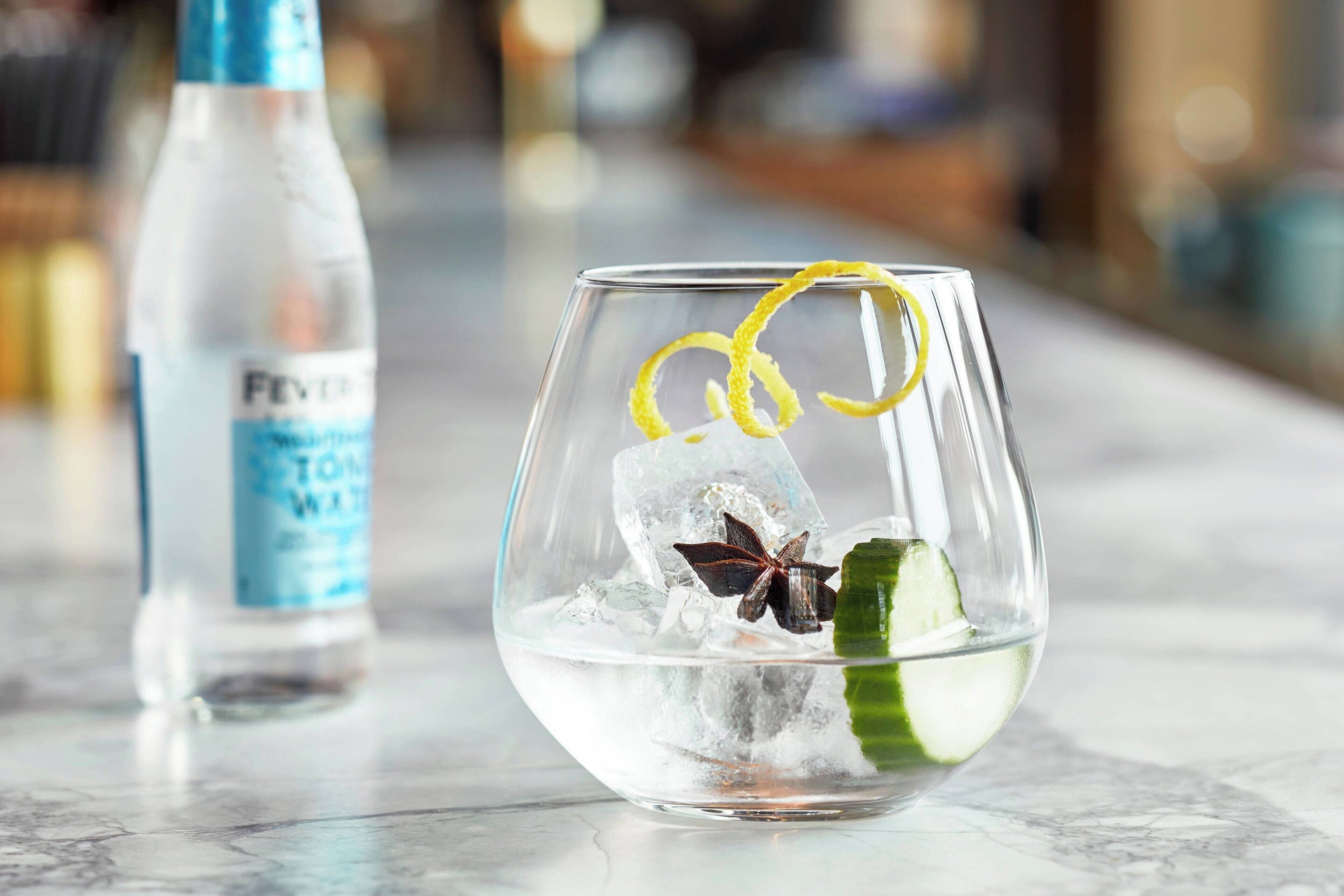 A clear glass with ice cubes, a star anise, and a lemon peel garnish on a marble surface.