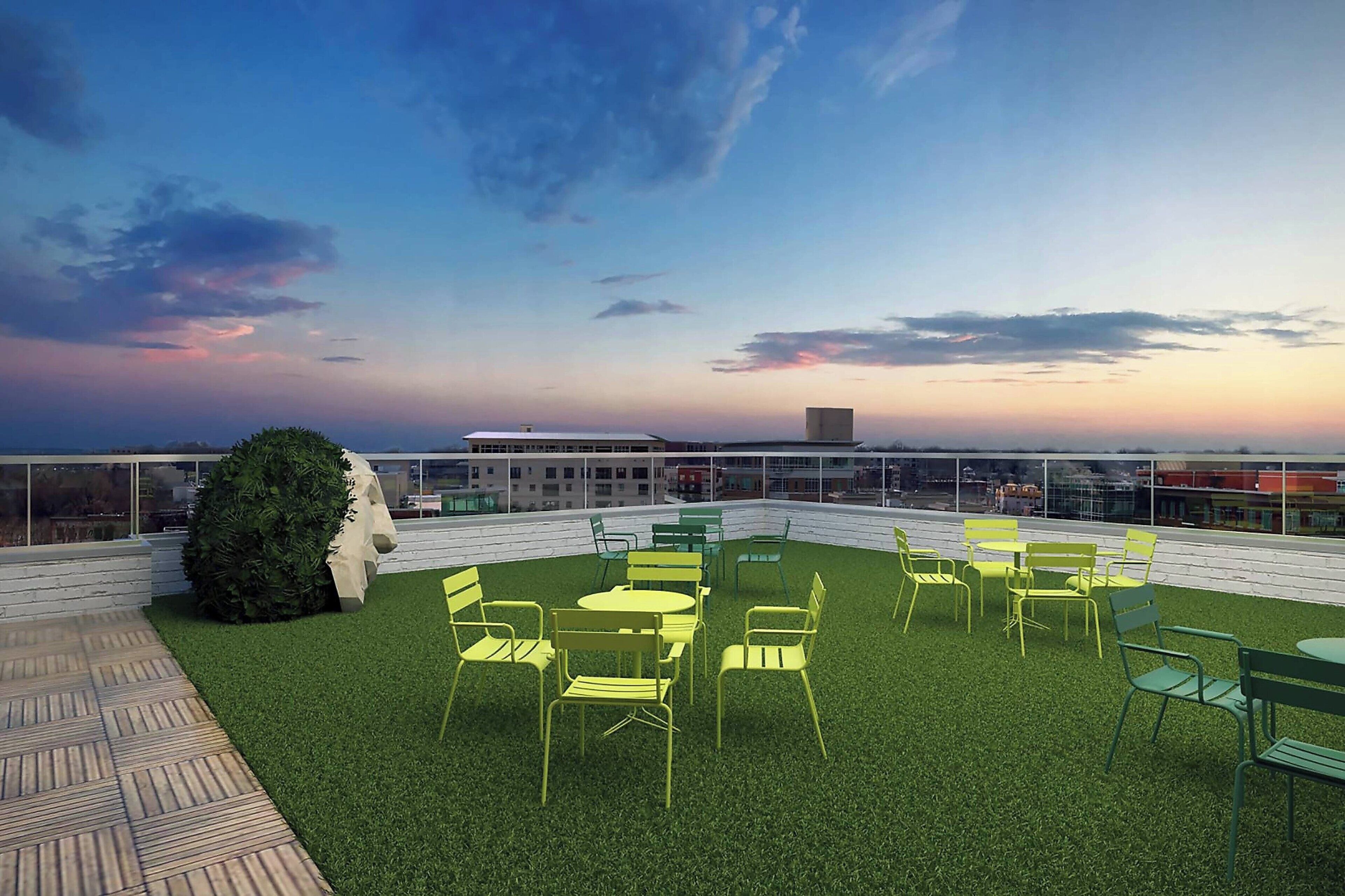A rooftop terrace with artificial grass, yellow and green chairs, and a view of the city skyline at dusk.