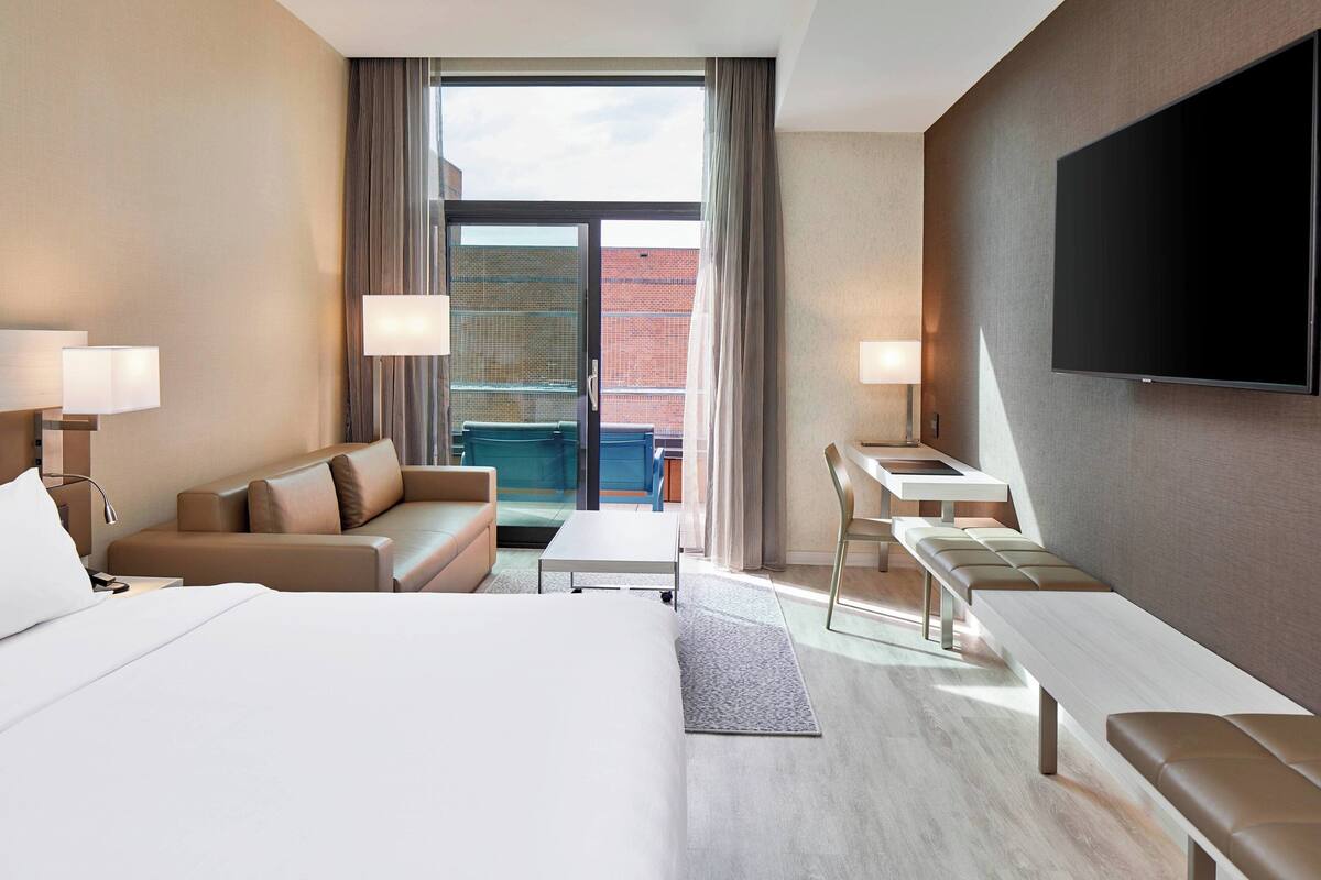 Une chambre d’hôtel avec un grand lit, un canapé, un bureau et une télévision. Elle dispose d’un balcon avec vue sur un immeuble.