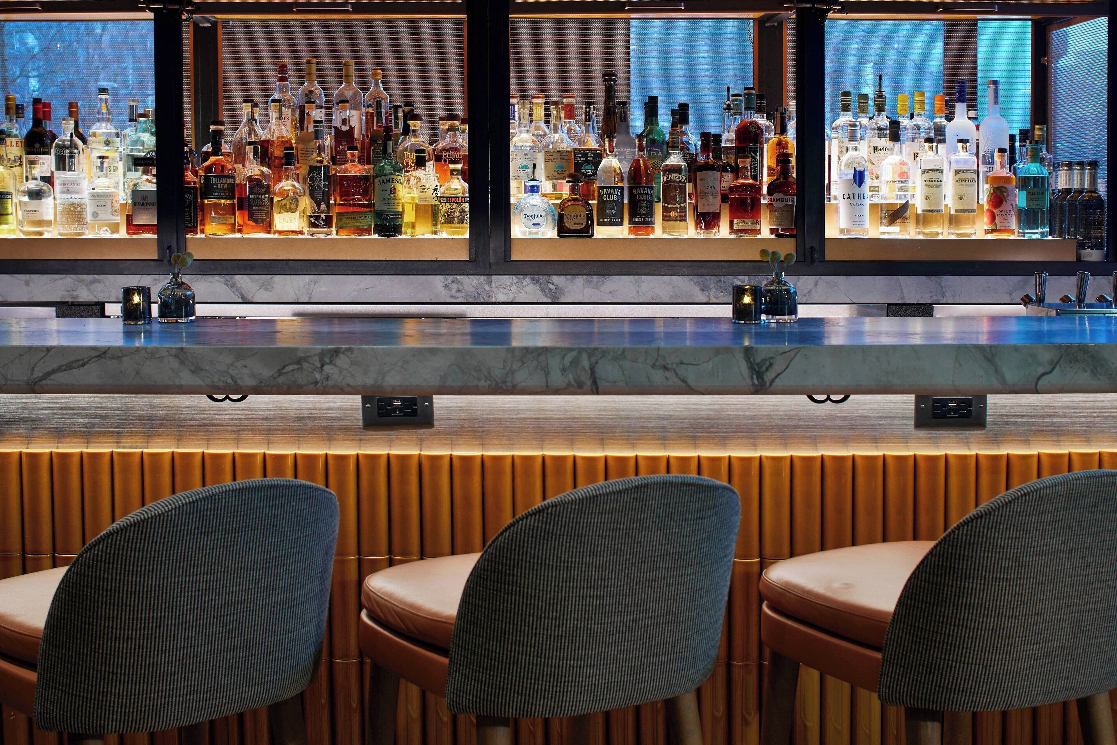 Een bar met verschillende drankflessen, een marmeren aanrecht en beklede barkrukken.