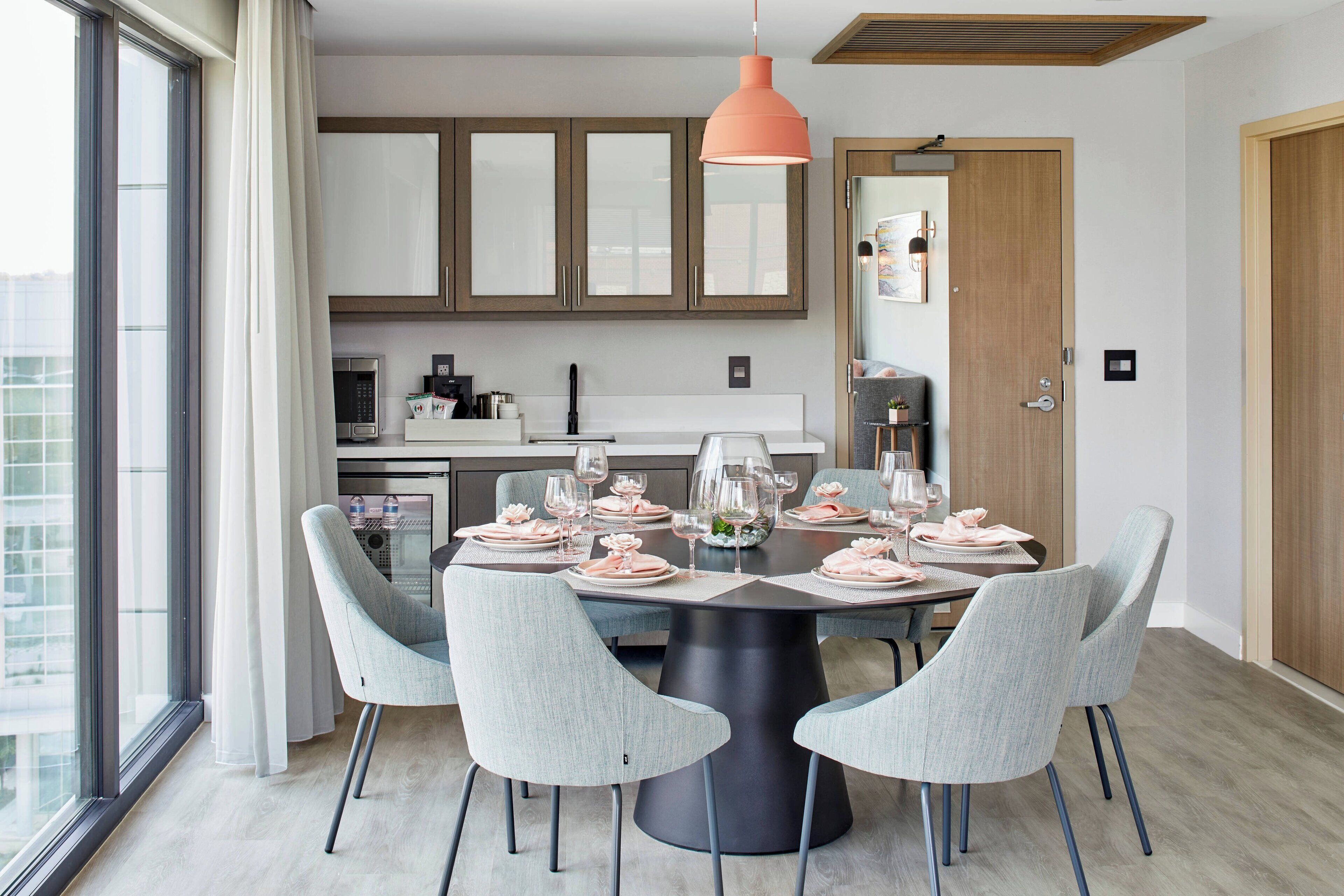 Un comedor moderno con una mesa redonda dispuesta para seis comensales, sillas tapizadas en gris claro y una lámpara colgante central.