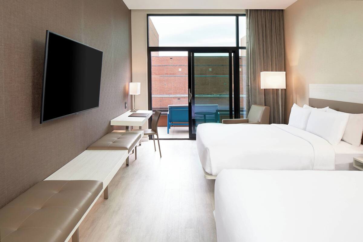 Une chambre d’hôtel dotée d’une grande télévision à écran plat, d’un lit, d’un bureau avec une chaise, d’un banc et offrant une vue sur l’extérieur grâce à une grande fenêtre.