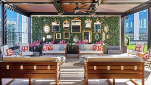 A modern lounge area with a green wall adorned with mirrors and flowers, wooden benches, and a ceiling fan.