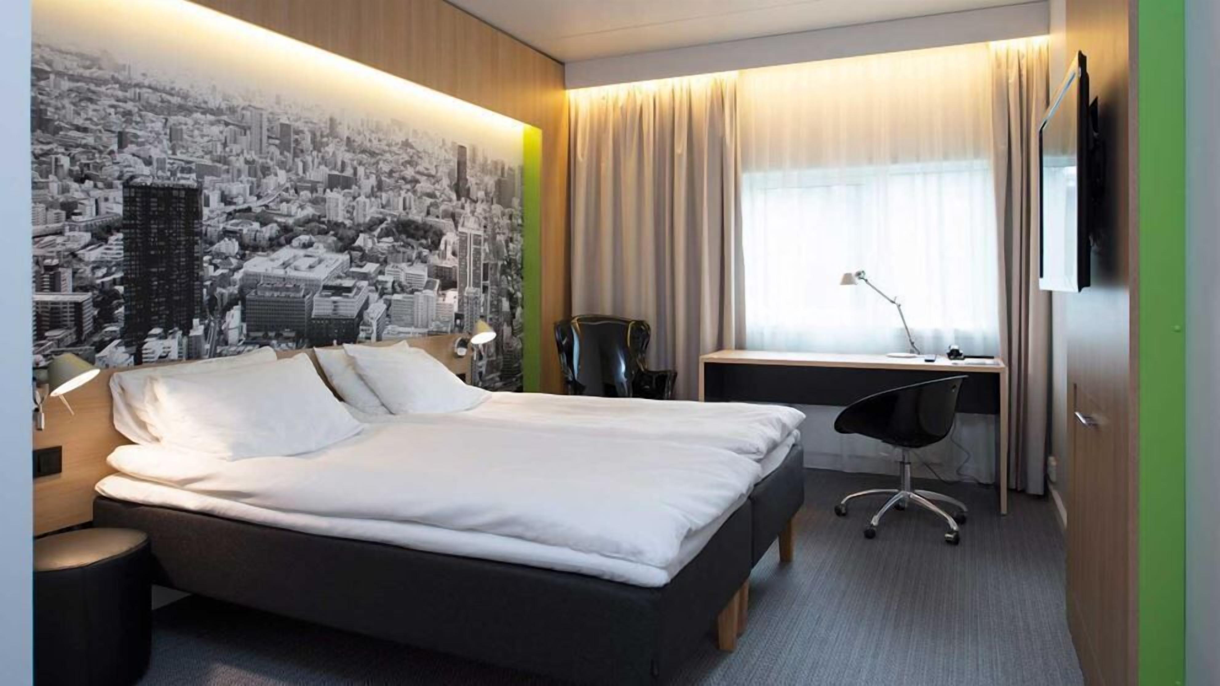 Habitación de hotel con cama, escritorio, silla y un mural de un paisaje urbano en la pared.