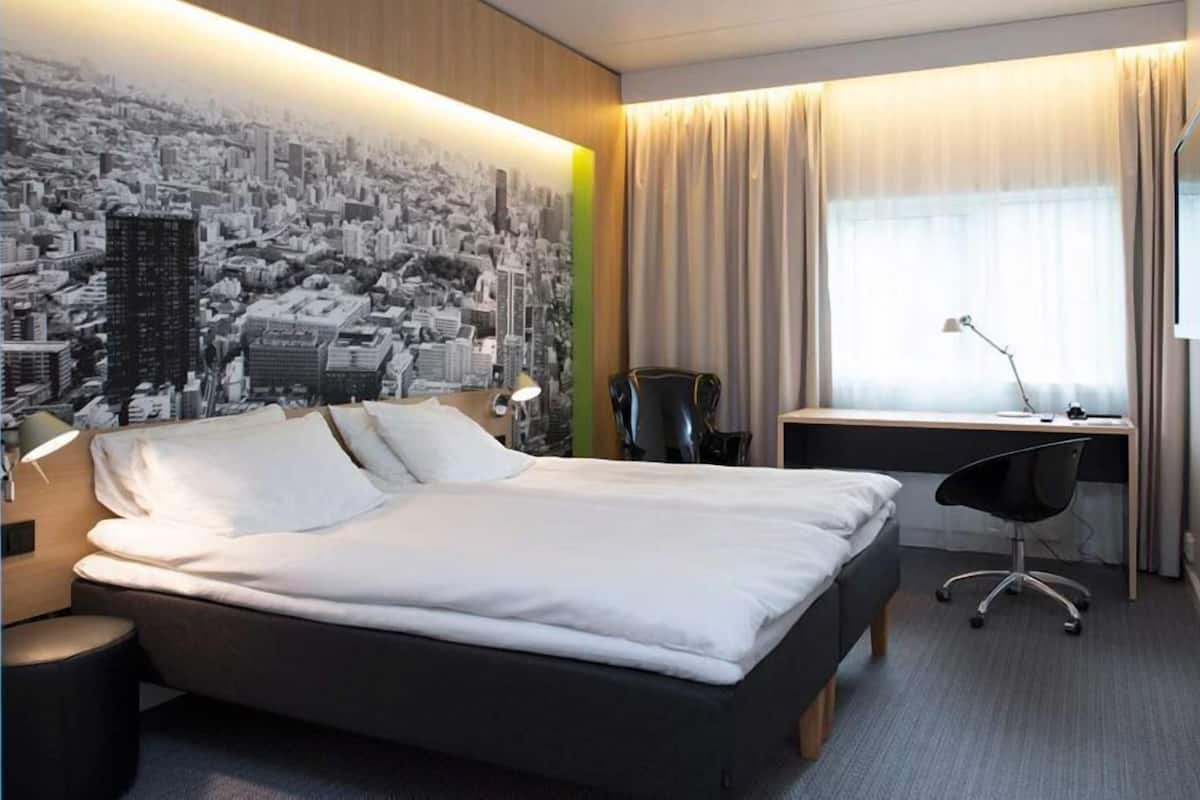 Habitación de hotel con cama, escritorio, silla y un mural de un paisaje urbano en la pared.