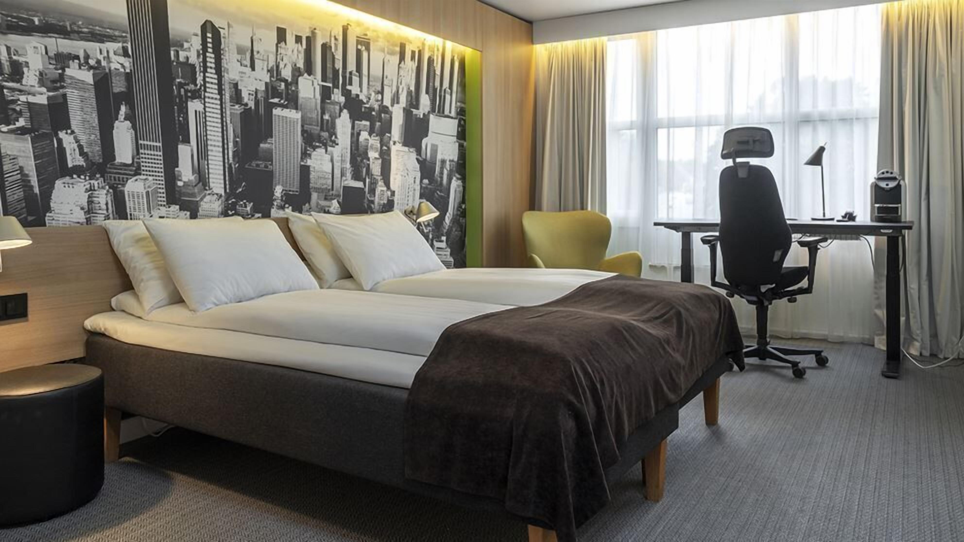 Habitación de hotel con una cama grande, un escritorio, una silla y un mural de un paisaje urbano en la pared.