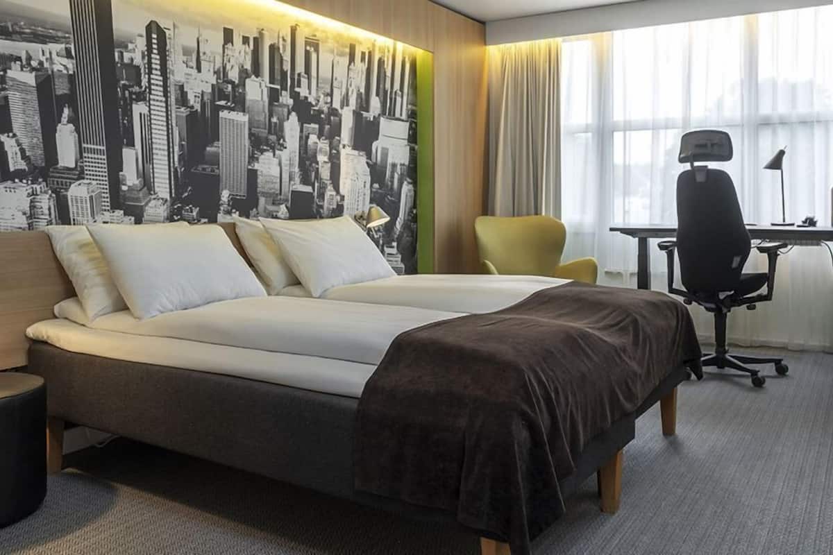 Habitación de hotel con una cama grande, un escritorio, una silla y un mural de un paisaje urbano en la pared.