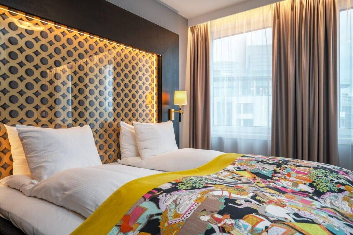 Una habitación de hotel con un cabecero estampado, una cama con sábanas blancas, una colcha colorida y una ventana con cortinas.