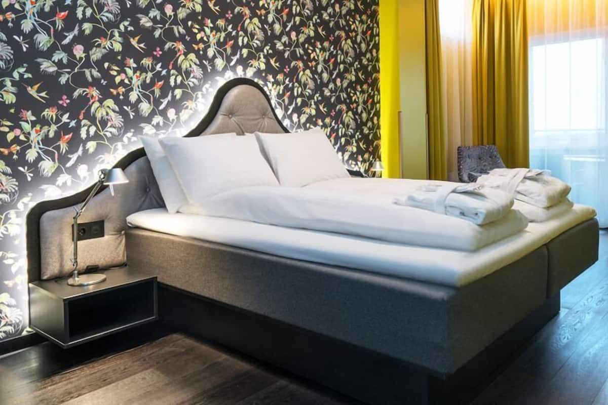 Habitación de hotel moderna con una cama grande, una mesita de noche con lámpara, una silla y vistas al exterior a través de una ventana.