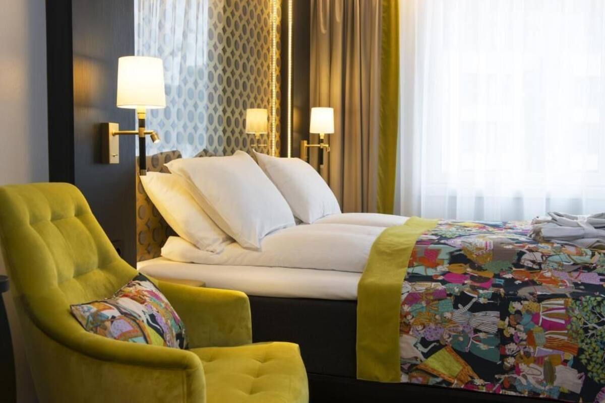 Habitación de hotel con cama, un sillón verde, dos lámparas de pared y una colcha estampada.