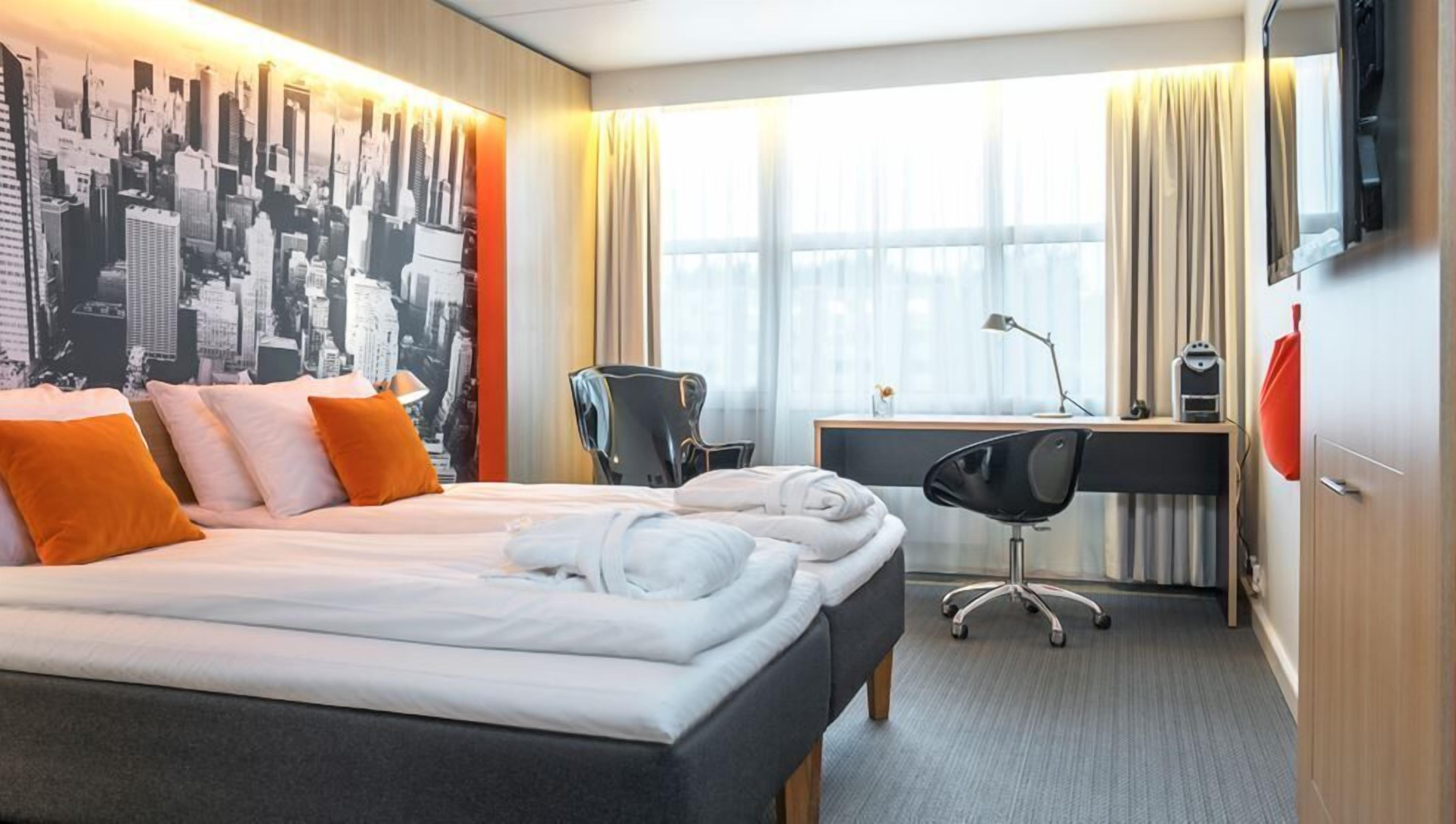 Habitación de hotel con cama, escritorio, silla, televisor y un mural con vista a la ciudad.