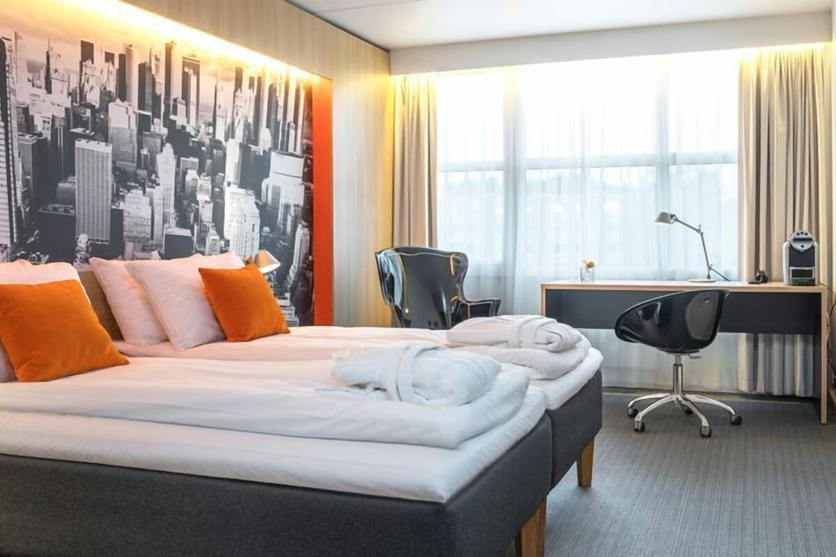 Habitación de hotel con cama, escritorio, silla, televisor y un mural con vista a la ciudad.