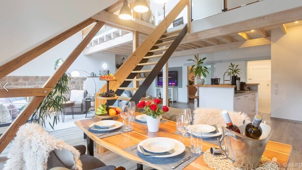 A dining area with a wooden table set for four, a fruit and cheese board, and a view of the kitchen and living area through an open space. - Holiday Apartment "Kleine Auszeit" – Your Place to Relax (Mürlenbach)