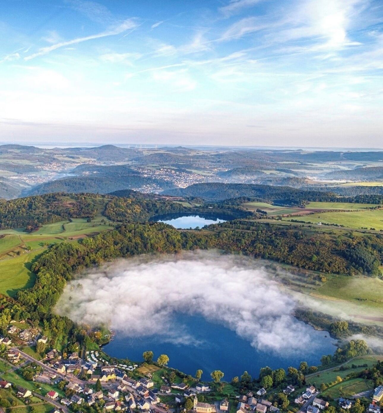 A lake surrounded by forested hills, a small village, and distant wind turbines.